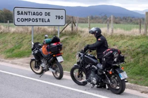 Dos motociclistas con maletas y conchas de peregrino posan con sus motos frente al gran cartel de entrada a la ciudad de Santiago de Compostela.
