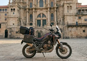 An adventure motorcycle loaded with travel panniers, parked in the center of Santiago de Compostela's Obradoiro Square, in front of the imposing baroque facade of the Cathedral.